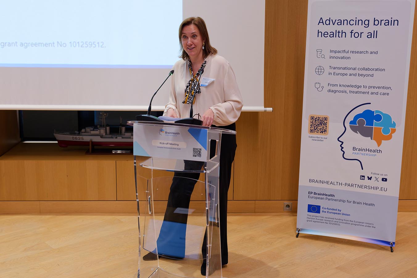 Person standing behind a clear lectern with a microphone in a conference room next to a banner about brain health