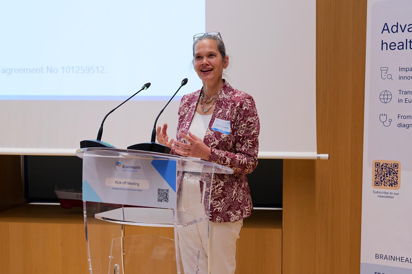 Woman with glasses on her head speaking at a clear podium with two microphones in a conference room in front of a banner about brain health