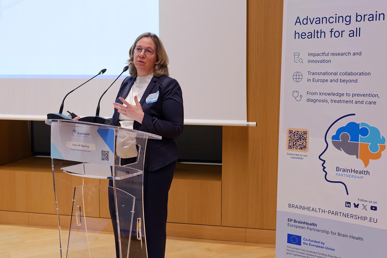 Person standing behind a transparent lectern speaking with a microphone in front of a banner about brain health