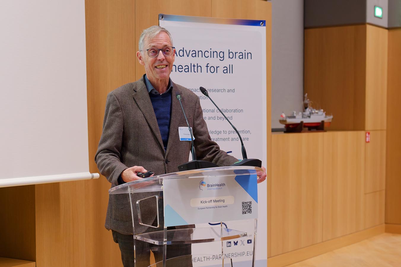Person standing behind a transparent lectern speaking with a microphone in front of a banner about brain health
