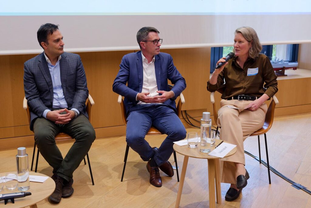 Three people sitting on chairs in a panel discussion setting with a small round table holding water bottles and glasses