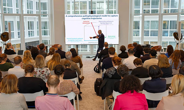Speaker pointing at a presentation slide in a conference room filled with seated attendees facing large windows
