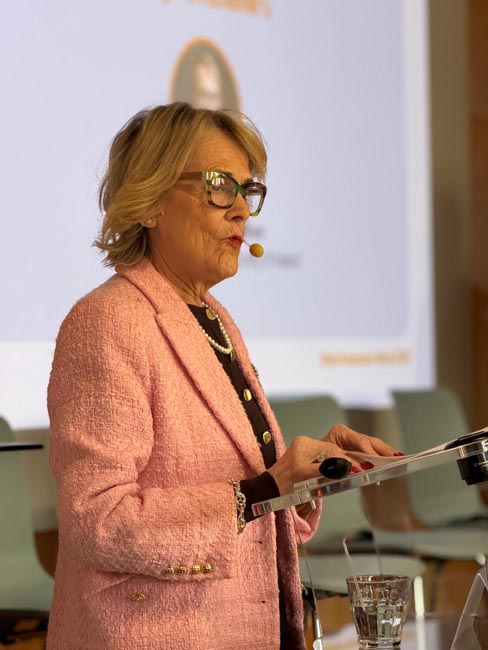 Woman in a pink textured blazer standing at a clear lectern with a microphone and glass of water in a conference room