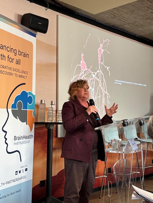 Person holding a microphone and speaking in front of a presentation screen showing a neuron diagram and a banner with a brain logo and text about brain health partnership