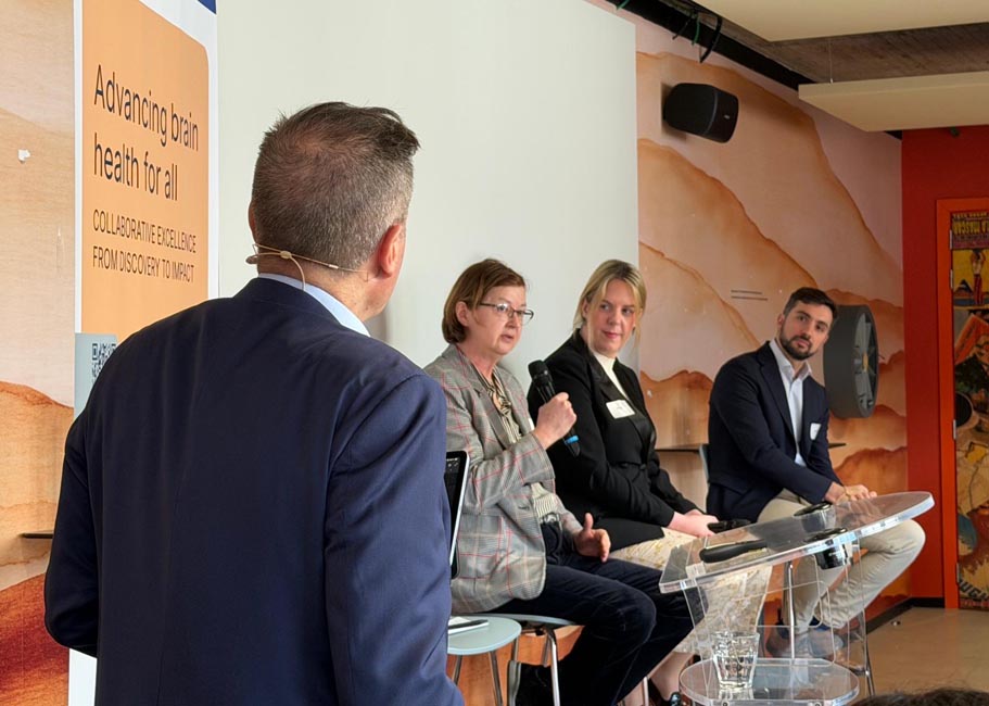 Four people in a discussion panel with one man standing and three seated behind a transparent lectern in front of a wall with a poster reading 'Advancing brain health for all'