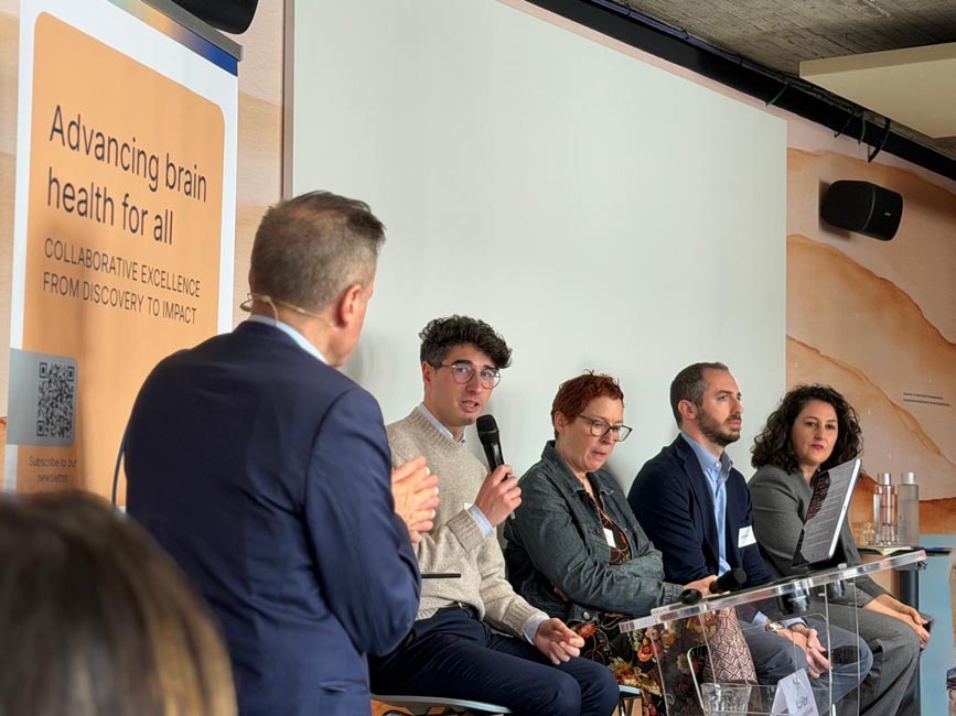 Panel of four people sitting behind a transparent lectern with one person speaking into a microphone and another person facing them in the foreground