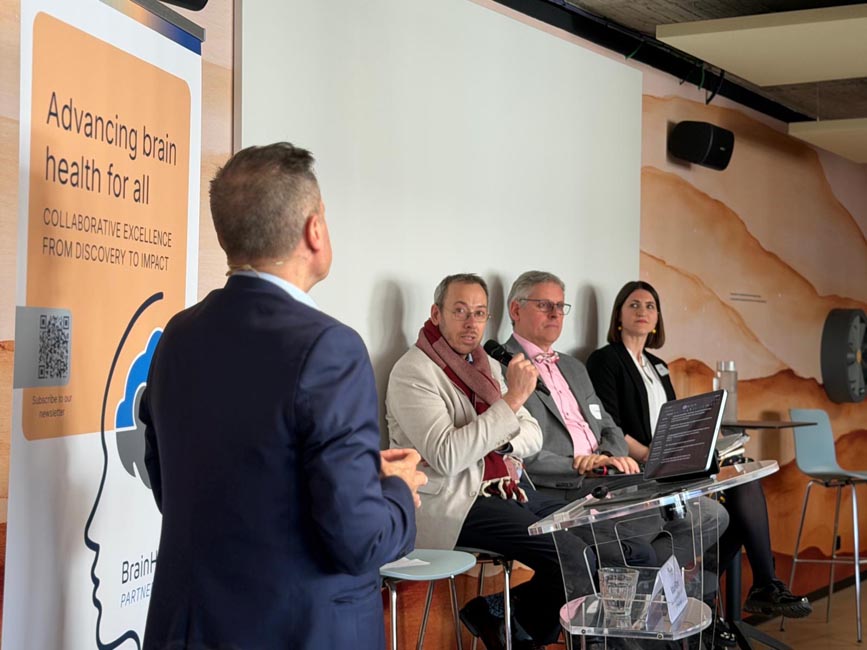 Man in suit speaking to three seated panelists at a conference table with a laptop and microphone