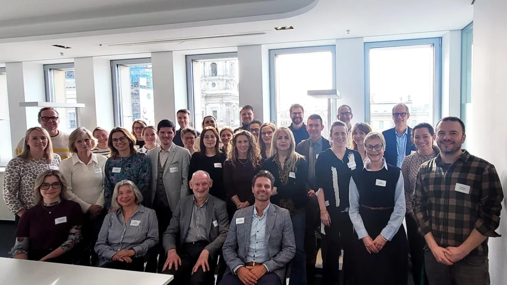 Group of people posing for a photo in a bright room with large windows showing a cityscape outside