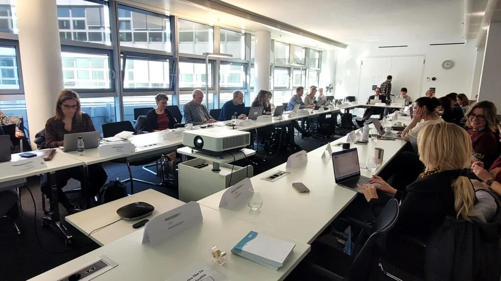 People sitting around a large conference table with laptops and papers in a bright room with windows